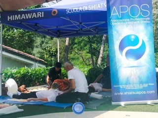 A group of people do Shiatsu treatments under a tent, next to an Apos banner.