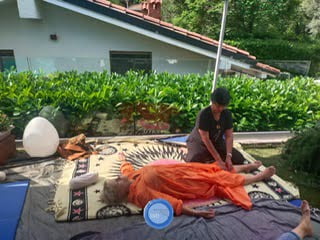 Un uomo con una veste arancione riceve un trattamento Shiatsu su una coperta davanti a una casa.