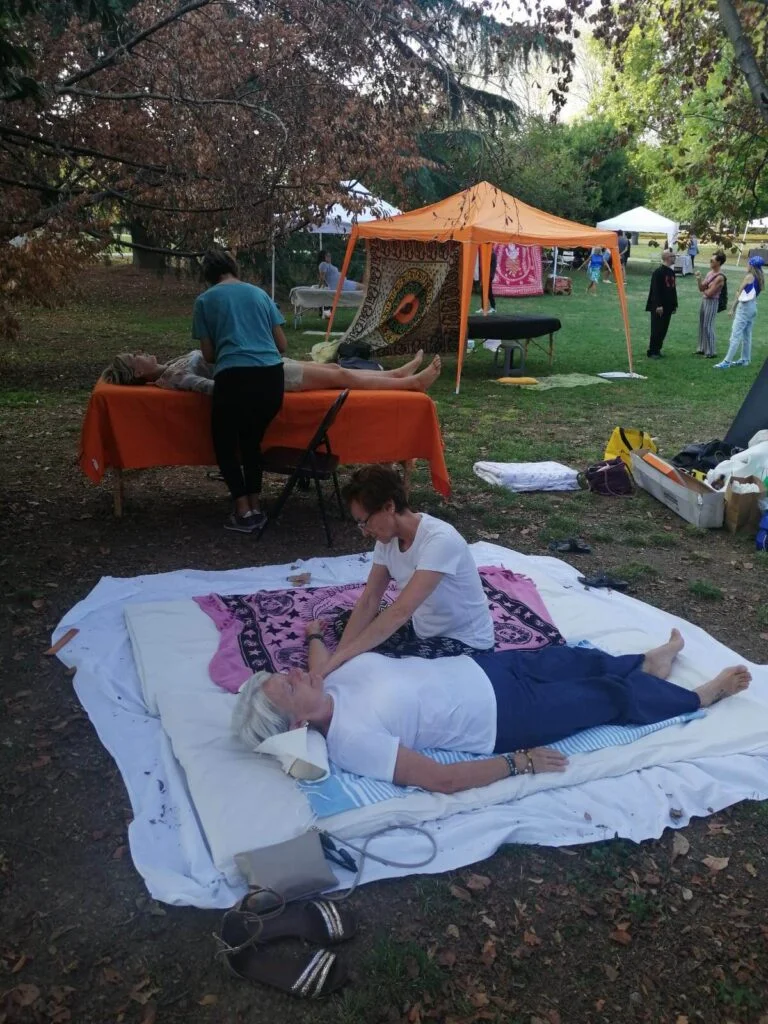 Inside a park, in the foreground treatment of the arm with a supine uke, in the background treatment of the abdomen with a supine uke on an orange bed, in the background various Shiatsu stations and gazebos.