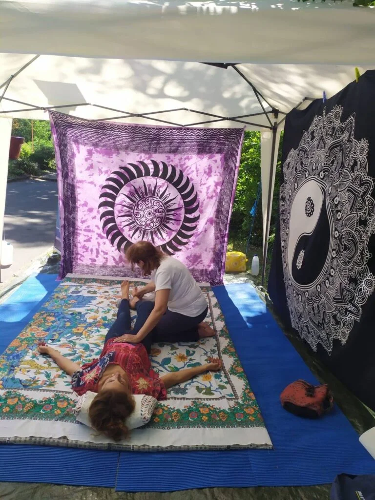 Leg treatment with mother hand on tanden on a stand set up in a public park with a large gazebo, mat and two sarongs, one black on a purple background and one white on a black background with oriental motifs (mandala, sun, tao symbol) used as walls against the sun
