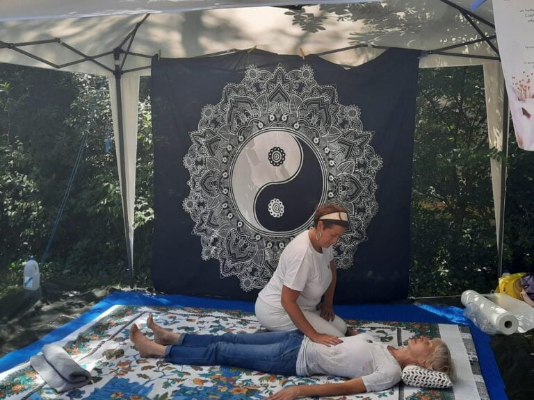 Treatment of the abdomen on a stand set up in a public park with a large gazebo, mat and two sarongs, one black on a purple background and one white on a black background with oriental motifs (mandala, sun, Tao symbol) used as walls against the sun at the his left can be seen roll up Apos
