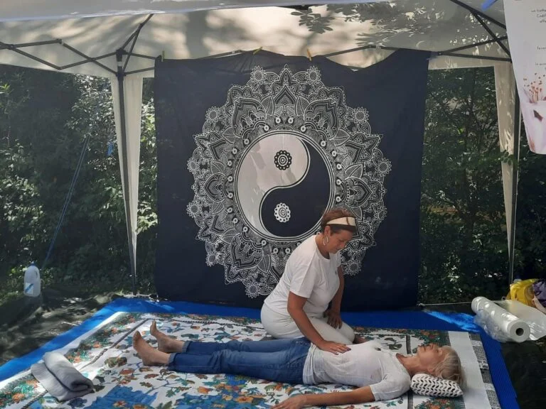 Treatment of the abdomen on a stand set up in a public park with a large gazebo, mat and two sarongs, one black on a purple background and one white on a black background with oriental motifs (mandala, sun, symbol of the tao) used as walls against the sun at on his left you can see a roll up Apos