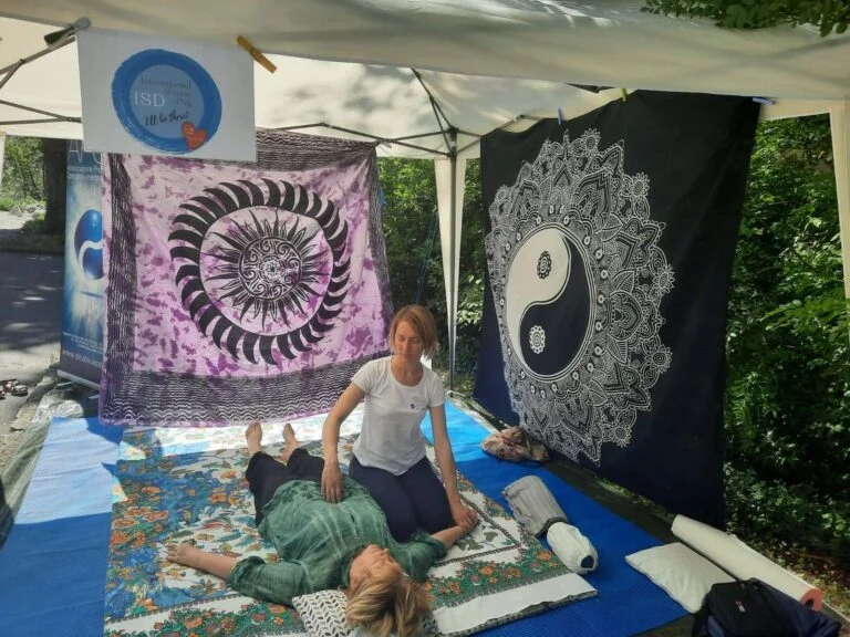 Treatment of the abdomen on a stand set up in a public park with a large gazebo, mat and two sarongs, one black on a purple background and one white on a black background with oriental motifs (mandala, sun, symbol of the tao) used as walls against the sun at on his left you can see a roll up Apos