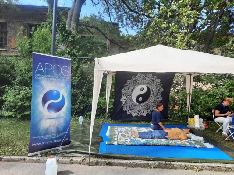 Treatment of the abdomen on a stand set up in a public park with a large gazebo, mat and white sarong on a black background depicting a mandala with the Tao symbol inside, to its left roll up Apos